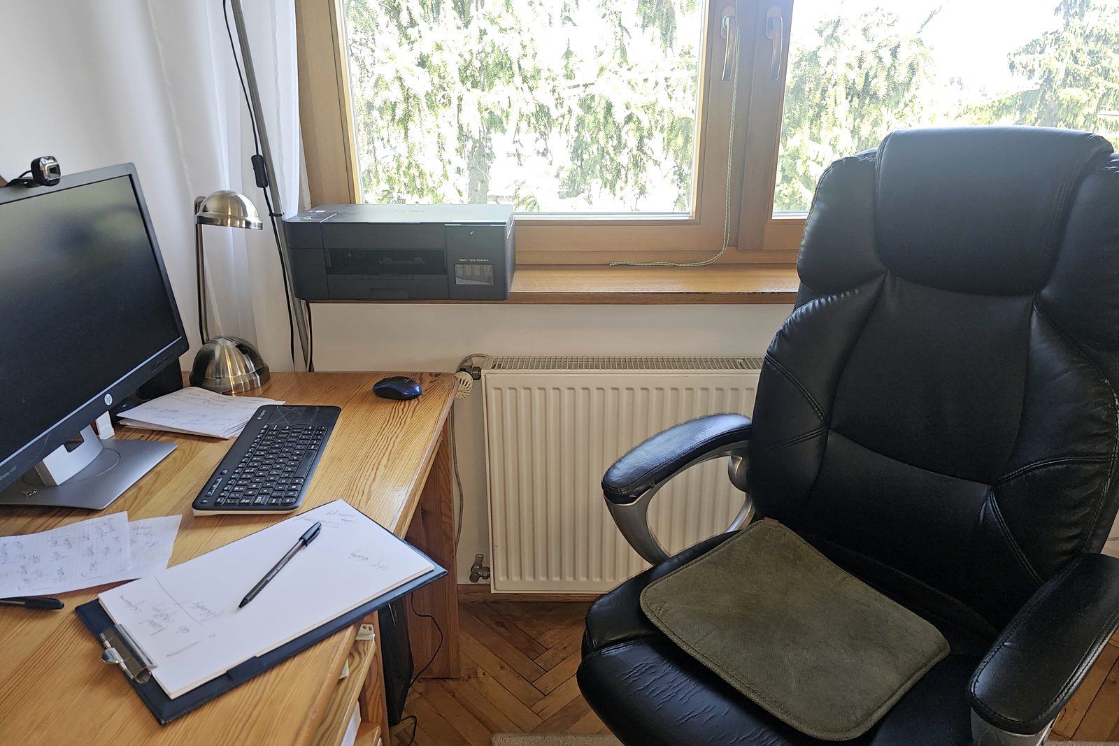 Biurko z monitorem i notatnikiem przy oknie z widokiem na drzewa, obok fotel